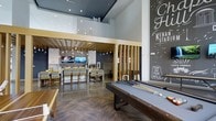 Located at 201 S Elliot Rd in Chapel Hill, NC, this vibrant single-floor community clubhouse is designed to foster social connection and leisure. The space makes a striking first impression with soaring double-height ceilings and an open-plan layout that seamlessly blends entertainment and gathering areas.

The game zone anchors one side of the room, featuring a full-size billiards table and a shuffleboard table set atop richly patterned herringbone-style flooring. Warm wood vertical slat dividers elegantly define zones without closing off the open feel of the space.

Adjacent to the game area, a communal dining and lounge section offers long bar-height tables with upholstered seating for groups, illuminated by a cascade of Edison-style pendant lights. A mosaic tile feature wall backs dual wall-mounted flat-screen televisions, creating an ideal setting for watching events together.

The design palette blends navy blue, natural wood tones, and polished concrete-style floors, lending the space a modern yet welcoming character. A bold chalkboard-style mural celebrating Chapel Hill landmarks — including references to Kenan Stadium and "Top of the Hill" — adds a strong sense of local identity and community pride throughout the space.