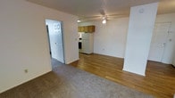 This single-floor unit presents an open-concept layout that connects the living area and kitchen in a practical, easy-to-navigate design. The living space is finished with wall-to-wall carpet, offering a comfortable underfoot feel, while the kitchen and adjoining area transition to wood-look hard flooring, providing a clean contrast between zones.

The kitchen is equipped with warm-toned oak cabinetry mounted above the counters, a white refrigerator, and a range, giving it a straightforward and functional character. A ceiling fan is centrally positioned overhead, serving both the kitchen and the open living space, contributing to air circulation throughout.

The walls are finished in a neutral white tone throughout, complemented by a textured ceiling that adds subtle visual interest. A six-panel interior door is visible leading to an additional area of the unit, suggesting further rooms beyond the main living space. White trim and baseboards frame the space consistently.

Overall, the unit offers a clean, modest, and functional layout well-suited for everyday living, with a simple, no-frills aesthetic that is easy to personalize.