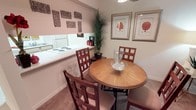 This single-floor space offers a comfortable and well-appointed living environment with a cohesive, welcoming aesthetic throughout. The dining area serves as a central feature, accommodating a round wood-finished table surrounded by richly stained lattice-back chairs, creating an intimate setting ideal for everyday meals or casual gatherings. A decorative bowl centerpiece adds a refined finishing touch to the dining setup. The space transitions seamlessly into the kitchen, which is accessible via an open pass-through counter, allowing for easy interaction between cooking and dining areas. The kitchen features a white electric cooktop and ample counter space, maintaining a clean and functional design. Warm, neutral tones dominate the walls, complemented by soft carpeting underfoot that adds to the overall sense of comfort. Decorative wall art, including framed botanical prints and textured square wall hangings, contributes a curated, stylish ambiance. A tall potted palm plant introduces a touch of natural greenery, enhancing the brightness and warmth of the interior. Recessed lighting overhead ensures the space is well-lit, while the overall layout and décor combine to create a functional yet inviting environment suitable for everyday living.