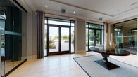 This elegantly appointed single-floor clubhouse facility offers a seamless blend of refined architectural design and upscale interior styling. The welcoming entrance lobby features large floor-to-ceiling glass doors and windows framed in dark metal, allowing abundant natural light to flood the light-tiled floors while offering views of the manicured exterior landscaping.

Inside, the main lounge area is a showpiece, boasting soaring double-height ceilings, a dramatic open staircase with sleek metal railings, and a striking sculptural chandelier. Comfortable leather and upholstered seating arrangements invite relaxation, while large glass doors provide direct views of an outdoor pool area.

A dedicated business center offers workstation computers arranged around a central table, bathed in natural light from tall windows framed by sheer drapery and a contemporary sputnik-style pendant fixture. An adjacent lounge area provides comfortable seating for informal gatherings.

One of the most distinctive spaces is a richly designed sitting room adorned with deep teal-painted wainscoting and coffered wall paneling, lending an intimate, club-like atmosphere. Tall ceilings, floor-length drapery, and warm wood accents complement the plush furnishings.

Additional lounge areas display vibrant artwork, crystal pendant lighting, and refined transitional furnishings, creating an overall ambiance that is both sophisticated and inviting throughout.