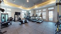 This fitness center is thoughtfully designed to support a comprehensive workout experience within a single, open floor. The space features a distinctive tray ceiling with warm ambient LED lighting and recessed fixtures, lending the room a polished, upscale atmosphere. Large windows fitted with horizontal blinds line the walls, allowing ample natural light to fill the space while maintaining privacy.

The cardio zone is well-stocked with multiple treadmills, elliptical trainers, and a stair climber, each equipped with individual monitors for entertainment during workouts. A wall-mounted flat-screen television adds to the cardio area's appeal. A spin bike is also available for cycling enthusiasts.

The strength training section features Matrix-branded cable and weight machines alongside a multi-functional rack system. A full dumbbell rack with a wide range of weights caters to free weight training needs. Functional fitness accessories — including kettlebells, medicine balls, resistance bands, TRX suspension trainers, a battle rope, and stability balls — are neatly organized on a dedicated storage rack.

Durable rubber tile flooring covers the entire space, providing cushioning and traction suitable for all types of exercise. A storage cabinet offers additional organizational convenience. The overall ambiance is clean, bright, and professional, making this fitness center a fully capable facility for diverse training goals.