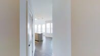 This single-floor residence at Overture showcases a thoughtfully designed open-concept layout that seamlessly connects the entryway, kitchen, and living areas. The space is immediately distinguished by its dramatically high ceilings, which rise well above standard height and are lined with a row of clerestory windows that flood the interior with natural light from multiple angles. Large windows along the perimeter walls further enhance the bright, airy atmosphere.

The kitchen serves as the focal point of the main living area, anchored by a generously sized island featuring a double sink, stone countertops, and warm wood cabinetry. Stylish pendant lights hang above the island, complementing a ceiling fan and additional track lighting overhead. Stainless steel appliances complete the modern kitchen aesthetic.

Dark hardwood-style flooring runs throughout the space, providing a rich contrast to the crisp white walls and bright ceiling. A louvered door near the entry suggests convenient storage or utility access. The open sightlines from the entry corridor to the living area create a welcoming sense of depth and spaciousness, making this a bright and functional modern residence.