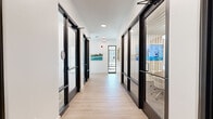 Located at 1301 Lebanon Pike in Nashville, Tennessee, this single-floor commercial hallway serves as the central circulation spine of a contemporary office environment. The space is thoughtfully designed with a crisp, modern aesthetic characterized by bright white walls, light wood-look flooring, and clean architectural lines that create a polished and professional atmosphere.

Flanking both sides of the corridor are floor-to-ceiling glass partition walls framed in bold black steel, offering visibility into adjacent private offices and a small conference room equipped with a wall-mounted display, a round table, and seating. The use of glass throughout promotes an open, transparent feel while maintaining functional separation between spaces.

A built-in white cabinetry station with a shiplap tile backsplash is integrated into one side of the hallway, providing practical storage and counter workspace. Framed artwork featuring natural landscapes is displayed along the walls, adding warmth and a touch of creativity to the otherwise minimalist environment.

Recessed ceiling lighting and flush-mount fixtures ensure consistent illumination throughout, while a glass-paned exterior door at the far end of the corridor allows natural light to filter in. The overall design balances functionality with aesthetic refinement, making it a welcoming and efficient transitional space within the commercial building.