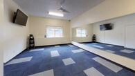 This single-floor fitness studio is designed as a functional and well-equipped exercise space, offering an open layout ideal for group classes or individual training sessions. The room features warm cream-colored walls complemented by a bold patterned carpet in a mix of blue and grey tones, adding visual interest to the floor while providing a comfortable surface underfoot.

A large wall-to-wall mirror spans one side of the room, enhancing the sense of space and allowing users to monitor their form during workouts. A wall-mounted flat-screen television is positioned at an elevated height, visible from across the room. Ceiling-mounted fluorescent lighting, along with a ceiling fan, ensures the space remains well-lit and ventilated during use. Multiple windows along one wall allow natural light to filter in, contributing to an energizing atmosphere.

Equipment is neatly organized along the perimeter of the room. A rack of kettlebells is positioned near the mirrored wall, while a dedicated storage area near the entrance houses stability balls in varying sizes, medicine balls, resistance bands, and exercise mats, all arranged on purpose-built storage racks. A frosted-glass door provides access to an adjacent area, completing this practical and versatile fitness space.