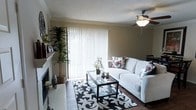 This two-floor residence offers a thoughtfully arranged living space with warm, neutral tones throughout. The living room serves as the heart of the home, featuring a classic brick fireplace along one wall and large sliding glass doors dressed with vertical blinds that allow abundant natural light to pour in. A ceiling fan with an integrated light fixture keeps the room comfortable year-round. Dark hardwood flooring anchors the space, complemented by a bold black-and-white patterned area rug that adds a striking visual contrast beneath a glass-top coffee table. The white sofa, accented with floral throw pillows, creates an inviting seating area, while tall indoor plants add a touch of greenery and warmth to the corners of the room. Crown molding along the upper walls adds a refined architectural detail, and a textured ceiling contributes additional character. The open layout allows for a natural flow between the living and dining areas, visible in the background. The overall ambiance is cozy yet polished, blending classic design elements with accessible, everyday comfort across both floors.