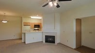 This single-floor space is designed with an open-concept layout that seamlessly connects the living area and kitchen. The living room is anchored by a built-in fireplace framed in light-toned tile, with a white surround that extends to the ceiling, creating a clean and prominent focal point. A ceiling fan with an integrated light fixture is centrally positioned overhead, providing both air circulation and ambient lighting. The flooring throughout the living area consists of neutral-toned carpet, contributing to a warm and comfortable atmosphere.

The kitchen is positioned adjacent to the living space and is accessible via a peninsula-style breakfast bar with a light countertop, offering additional seating and workspace. The kitchen features light wood cabinetry, black built-in appliances including a microwave and stovetop, and a white refrigerator. An overhead fluorescent light fixture provides direct task lighting above the kitchen workspace. The neutral color palette used throughout—from the white walls to the light cabinetry—creates a cohesive, understated aesthetic that offers a versatile backdrop for a variety of interior styles.