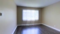 This single-floor space offers a clean and functional layout with understated, practical design. The interior is finished with rich, dark hardwood flooring that runs throughout, adding warmth and a polished look to the space. The walls are painted in a neutral warm beige tone, creating a calm and welcoming atmosphere that pairs well with natural light. A standout feature of the main room is the large picture window dressed with vertical blinds, which allows generous natural light to pour in, casting bright streaks across the hardwood floor. The window offers a pleasant view of lush greenery outside, connecting the interior with the natural surroundings. White baseboards provide a crisp contrast against both the flooring and the wall tones, contributing to a tidy, well-maintained appearance. The room is open and uncluttered, lending itself well to a variety of furniture arrangements and uses. The overall ambiance is bright, airy, and move-in ready, making it a versatile space suitable for comfortable everyday living.