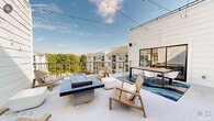 The Sky Lounge in Wake Forest, North Carolina is a thoughtfully designed two-floor rooftop amenity space that offers an elevated outdoor retreat. The open-air terrace is finished with a clean white deck surface and features string lights overhead, creating an inviting atmosphere from day into evening. The main rooftop level is organized into distinct gathering zones, including a fire pit area flanked by teak-style lounge chairs and a mounted outdoor television, ideal for relaxed social gatherings. A dedicated outdoor dining area with a large table and modern white chairs sits atop a vibrant blue wave-patterned area rug, providing a comfortable setting for al fresco meals. The space is framed by sleek black metal railings that open up to sweeping views of the surrounding tree canopy and residential buildings. A covered lounge area offers shade and shelter, outfitted with a generous sectional sofa accented with colorful throw pillows, a concrete coffee table, and a ceiling fan overhead — all set against crisp white shiplap walls. The overall aesthetic is coastal-contemporary, combining natural wood tones, neutral upholstery, and clean architectural lines. The Sky Lounge delivers a versatile, well-appointed outdoor experience with a relaxed yet refined ambiance.