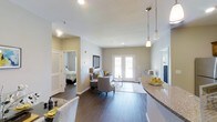 This well-appointed single-floor residence offers an open-concept design that seamlessly connects the kitchen, dining, and living areas into one cohesive, light-filled space. The kitchen features granite countertops and pendant lighting that hang elegantly above the breakfast bar, providing both functional workspace and casual seating. Rich, dark hardwood flooring runs throughout the main living area, adding warmth and sophistication to the neutral-toned interior.

The living area is bright and welcoming, centered around a comfortable seating arrangement and framed by a pair of classic French doors that invite natural light and provide direct access to the outdoors. The smooth white ceilings and soft, neutral wall tones create a clean, airy ambiance throughout.

A glimpse into the bedroom reveals a spacious and serene retreat, complete with louvered closet doors that offer both style and ample storage. Recessed lighting throughout the unit ensures consistent, warm illumination in every area. The overall design balances functionality with comfort, making this a practical yet attractive living space suited for everyday life.