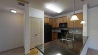 This single-floor space centers around a well-appointed kitchen designed for both practicality and style. The kitchen features warm-toned wood cabinetry extending to the upper walls, providing generous storage throughout. Dark granite countertops run along the perimeter and continue onto a peninsula, which includes a double stainless steel sink with a modern gooseneck faucet, creating a natural division between the kitchen and adjacent living areas. A pendant light hangs above the peninsula, complementing the flush-mounted fluorescent fixture overhead. Black appliances, including a refrigerator, microwave, and range, create a cohesive, contemporary aesthetic against a dark tile backsplash. The hardwood-style flooring adds warmth to the space. A set of double bi-fold doors nearby likely conceals a storage closet or laundry area, adding functional versatility. The layout makes efficient use of its footprint, with an open sightline through the kitchen that enhances the sense of flow. Overall, the space offers a practical and cohesive interior with consistent finishes throughout.