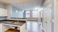 This single-floor unit is designed with an open, flowing layout that connects the kitchen and living areas seamlessly. The kitchen is well-appointed with white shaker-style cabinetry, dark granite countertops, and a full suite of stainless steel appliances, including a refrigerator, range, microwave, and dishwasher. A peninsula with an undermount sink and pendant lighting above defines the kitchen boundary while maintaining visual openness to the living space.

The living area extends generously from the kitchen, finished with warm hardwood-style flooring that runs throughout the unit. Soft blue-gray walls lend a calm, neutral tone to the interior. Large windows in the main living space admit abundant natural light and frame views of the surrounding urban streetscape.

The bedroom is a well-proportioned room with its own window for natural light and a ceiling fan overhead, continuing the hardwood-style flooring and coordinating wall tones.

The bathroom is equipped with an open vanity with a white countertop, a wide mirror, and a combination tub and shower. Grab bars are installed in the tub area, reflecting an accessibility-conscious design. The overall ambiance of the unit is clean, modern, and functional.