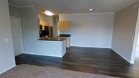 This single-floor residence offers a clean, modern design with an open-concept layout that maximizes both space and functionality. The kitchen is thoughtfully positioned as the centerpiece of the main living area, featuring a peninsula with a dark countertop that provides additional workspace and a natural divider between the kitchen and living space. Light wood cabinetry lines the kitchen walls, offering ample storage, while stainless steel appliances — including a refrigerator and additional built-in units — add a contemporary touch. Recessed lighting and a flush ceiling fixture keep the space well-lit throughout. Dark hardwood flooring flows through the main living and kitchen areas, creating a rich, grounded aesthetic, while a transition to carpet hints at a separate lounging or living zone. The neutral, light-toned walls throughout the space lend a bright and airy ambiance, making the interior feel open and inviting. The overall layout is practical and versatile, well-suited for everyday living, with a straightforward flow that connects each area naturally. A doorway visible near the kitchen suggests access to additional rooms, completing this well-appointed single-level home.