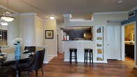 This single-floor residence showcases a thoughtfully designed open-concept layout that seamlessly connects the living and dining areas to the kitchen. Rich, dark hardwood floors run throughout the main living spaces, adding warmth and a sense of sophistication to the interior. Classic crown molding frames the ceilings and architectural columns, lending the space an elegant, refined character.

The kitchen is defined by a half-wall breakfast bar with a dark countertop, offering casual seating for two with stylish black bar stools. Dark cabinetry in the kitchen creates a striking contrast against the light-toned walls, while the pass-through design keeps the space open and connected to the rest of the living area. Built-in open shelving along one wall provides functional display space, currently accented with decorative books and curios.

The dining area accommodates a full table and chairs, positioned comfortably near the kitchen for easy entertaining. A framed art piece on the wall and carefully placed décor throughout contribute to the warm, inviting ambiance. A hallway visible in the background suggests additional private rooms beyond the main living space. Overall, the residence balances classic architectural details with a modern, functional design aesthetic.