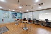 Located at 2037 Polo Gardens Drive in Wellington, Florida, this single-floor community amenity space offers a well-appointed and versatile environment for residents. The space is divided into two distinct functional areas that flow seamlessly into one another.

The business center section features a row of individual computer workstations separated by privacy partitions, providing a focused setting for work or study. Warm hardwood-style flooring runs throughout, complemented by a curved ceiling-mounted track lighting system that ensures bright, even illumination. A wall-mounted flat-screen television and colorful abstract artwork adorn the soft blue accent walls, adding a welcoming, modern touch.

Adjacent to the business center is a casual lounge and refreshment area, furnished with high-top bistro tables and bar-height chairs ideal for informal gatherings. A built-in coffee and beverage station with cabinetry provides convenient refreshments. This area connects directly to a grand lobby featuring soaring ceilings, expansive windows, and sliding glass doors that fill the space with abundant natural light and offer views of the outdoor surroundings. The lobby is furnished with contemporary sofas, accent chairs in vibrant teal and yellow tones, and stylish pendant lighting, creating an inviting and social atmosphere throughout.