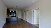 This single-floor space offers a thoughtfully designed open-concept layout that seamlessly connects the kitchen and living areas. The kitchen is a standout feature, showcasing dark espresso cabinetry, granite countertop surfaces, and a peninsula island that doubles as a casual dining or prep area. Pendant lighting hangs above the island, complemented by a track lighting system that adds both function and style to the cooking space. A black refrigerator anchors the kitchen, giving it a cohesive, contemporary aesthetic. Rich, dark hardwood flooring runs throughout the space, adding warmth and a sense of continuity between areas. One of the most eye-catching architectural details is the sliding barn door, mounted on a black metal track, which adds a modern-rustic character to the interior. Adjacent to the barn door is a standard door leading to additional rooms or closet space, providing practical storage solutions. The walls are finished in a neutral warm beige tone, creating a clean and inviting backdrop that complements the darker flooring and cabinetry. The smooth white ceiling and crisp white trim throughout the space lend a polished, finished look. Overall, the space combines contemporary design elements with functional living in a cohesive and well-appointed layout.