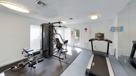This single-floor fitness center offers a practical and well-equipped space for a well-rounded workout experience. The room features light gray walls, a textured ceiling with flush-mount lighting, and a durable flooring surface with rubber matting placed beneath the equipment for safety and stability.

The facility is anchored by a multi-station cable weight machine, which accommodates a variety of strength-training exercises. Accompanying it is a flat weight bench, providing additional options for resistance training. For cardiovascular exercise, the space includes a treadmill and an elliptical trainer, both positioned to make efficient use of the room's layout.

Natural light enters through multiple windows fitted with horizontal blinds, as well as several glass-paneled exterior doors that also provide direct outdoor access. The doors allow views of surrounding greenery, contributing to a bright and open atmosphere. A fire extinguisher is mounted near one of the exits, and posted exercise room rules ensure safe usage of the space.

The overall design is clean, functional, and straightforward, prioritizing usability. The combination of cardio and strength equipment within a naturally lit, accessible environment makes this fitness center a convenient and complete exercise facility.