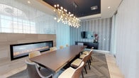 This single-floor space showcases a thoughtfully designed conference and dining area that blends modern elegance with functional sophistication. The room centers around a substantial dark wood rectangular table surrounded by cushioned chairs with cream upholstery, comfortably accommodating up to ten guests. Overhead, a dramatic multi-bulb brass chandelier serves as a bold focal point, casting warm ambient light throughout the space. A recessed tray ceiling painted in deep charcoal frames the chandelier, adding architectural depth and contrast to the room. Along one wall, a sleek linear gas fireplace framed in light wood veneer provides a warm, inviting atmosphere, while a mounted flat-screen television positioned against a textured, vertically striped accent wall offers multimedia functionality. A dark credenza beneath the television provides additional storage and display space, accented with decorative vases and floral arrangements. The flooring features a neutral-toned patterned carpet that complements the room's refined color palette of charcoal, taupe, and cream. Together, these elements create a polished, contemporary environment well-suited for meetings, gatherings, or formal dining.