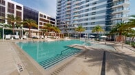 This expansive outdoor amenity space presents a striking resort-style pool deck designed to complement the contemporary high-rise tower that serves as its backdrop. The centerpiece is a generously sized swimming pool with crystal-clear turquoise water, framed by wide, elegant stone-tiled decking that invites relaxation and leisurely movement throughout the area. A spa or wading feature connects seamlessly to the main pool, adding a layered, resort-caliber experience to the amenity level. Rows of white lounge chairs and sun umbrellas line the pool perimeter, providing ample space for sunbathing and outdoor lounging. Lush tropical palm trees are thoughtfully interspersed throughout the deck, introducing a vibrant, resort-like atmosphere and natural shade. Pergola-style shade structures with slatted wood detailing offer additional covered retreats along the edges of the space. The surrounding high-rise tower rises dramatically overhead, featuring a modern façade of floor-to-ceiling glass windows and private balconies that overlook the pool below. The overall ambiance is clean, open, and sophisticated, blending contemporary architecture with a tropical outdoor aesthetic that evokes a seamless indoor-outdoor living experience.