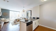 Located on Wilshire Boulevard in Los Angeles, this one-bedroom, one-bathroom residence offers a well-designed layout that maximizes every square foot of its compact footprint. The open-plan kitchen and living area forms the heart of the home, anchored by a sleek kitchen island with dark granite countertops, a stainless steel sink, and a full suite of stainless steel appliances, including a refrigerator, range, and built-in microwave. White cabinetry provides ample storage, while warm hardwood flooring ties the kitchen seamlessly into the living area. The living room is bathed in natural light from expansive floor-to-ceiling windows that frame urban views of the surrounding cityscape, creating a bright and airy atmosphere. An accent wall adds a contemporary touch to the living space. The bedroom is a quiet retreat, featuring plush carpeting, a large window with city views, and a dedicated workspace area, along with a closet for storage. The bathroom continues the clean aesthetic with hardwood-style flooring, a white vanity with under-sink storage, and a shower-tub combination. A hallway connects the rooms efficiently, completing this thoughtfully arranged urban residence.
