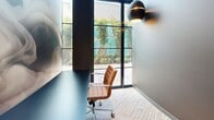 Located at 45 Northeast 27th Street in Miami's vibrant Wynwood neighborhood, this private office is a thoughtfully designed single-room workspace that makes a strong impression despite its compact footprint. The office is accessed through a sleek glass-paneled entry door framed in matte black, offering a preview of the refined interior beyond. Inside, the space features a striking full-wall photographic mural depicting soft, swirling smoke tones in neutral grays and whites, creating an artistic focal point that sets a creative and sophisticated atmosphere. A dark, minimalist desk is positioned facing a floor-to-ceiling window, which floods the room with natural light and frames a lush view of tropical greenery outside — a distinctly Miami touch. The carpeted floor features a textured woven pattern, adding warmth underfoot, while a sculptural globe pendant wall sconce in matte black provides ambient accent lighting. The glass partitions that define the office's walls allow visual connectivity to the surrounding environment while maintaining privacy through frosted panel detailing. The overall design blends modern minimalism with bold artistic expression, making this office a compelling and focused workspace in one of Miami's most dynamic districts.