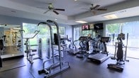Located at 8800 Pine Forest Rd in Pensacola, Florida, this single-floor fitness center is thoughtfully designed to support a well-rounded workout experience. The space is outfitted with a comprehensive selection of exercise equipment, including treadmills, elliptical machines, a stationary bike, and a multi-function cable and strength training tower, all arranged on durable rubberized flooring for safety and comfort. Large windows line the perimeter of the room, flooding the space with abundant natural light while offering pleasant views of the surrounding greenery and landscaped grounds outside. Two ceiling fans are mounted overhead, promoting consistent airflow and a comfortable environment during workouts. Wall-mounted flat-screen televisions provide entertainment options, while recessed lighting and a tray ceiling detail add a polished, finished look to the interior. The soft blue-gray walls contribute to a calm and focused atmosphere. Full-length mirrors are positioned along one wall to assist with form and technique. The overall design balances functionality with an inviting ambiance, making this a practical and well-equipped fitness facility for regular use.