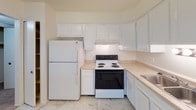 This single-floor residence offers a clean, functional layout designed for comfortable everyday living. The kitchen is well-equipped with white cabinetry extending to the ceiling for generous storage, light-toned countertops, a double-basin stainless steel sink, a dishwasher, and a full suite of white appliances including a refrigerator and electric range. The flooring features a marble-look tile that adds a bright, polished feel to the space. The open living area is generously sized, with plush wall-to-wall carpeting and a view into the kitchen through a pass-through opening, creating a connected and airy atmosphere. A sliding glass door brings in abundant natural light and looks out toward an outdoor greenspace. The bedrooms are well-proportioned, each finished with soft carpeting and large windows dressed with vertical blinds that allow for adjustable light control. The bathroom is tastefully finished with a marble-patterned vanity countertop, a wide mirror with strip lighting above, white cabinetry below, and a tiled tub-shower combination. Throughout the unit, neutral tones and consistent finishes create a cohesive, move-in-ready environment suited for a variety of lifestyles.