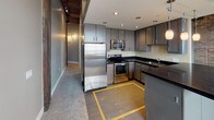 Located at 675–699 Lafayette St in Toledo, Ohio, this single-floor loft unit showcases a compelling blend of historic industrial architecture and contemporary interior design. The space is defined by its exposed brick walls, which span multiple rooms and serve as a striking visual anchor throughout, complemented by heavy timber beam ceilings that reinforce the building's warehouse heritage. Ductwork and track lighting are left intentionally visible, adding to the industrial aesthetic.

The open living area is generously proportioned, featuring plush neutral carpeting and large black-framed windows that frame views of the surrounding Toledo cityscape, allowing ample natural light to fill the space. A kitchen peninsula with a dark countertop separates the living area from the kitchen, which is equipped with stainless steel appliances—including a refrigerator, range, and built-in microwave—paired with gray cabinetry, dark granite countertops, and tile flooring with a decorative inset.

The carpeted bedrooms continue the brick and neutral-tone palette, with one room featuring an arched window overlooking the city. The bathroom includes a dark granite vanity, tile flooring, and clean modern fixtures. A ceiling fan, recessed lighting, and a rustic barn-style door add functional and decorative detail throughout.