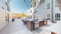 Located on North High Street in Columbus, Ohio, this thoughtfully designed outdoor courtyard serves as a central amenity space nestled within a modern multi-story residential building. The space is characterized by a generous composite deck that extends across the courtyard, framed by the building's contemporary gray siding and white paneled facades with private balconies overhead.

The courtyard is organized into several distinct functional zones. A dedicated grilling area features multiple stainless steel gas grills built into warm wood-clad outdoor kitchen stations, providing ample space for cooking and entertaining. Adjacent to the grilling area, a central dining zone offers a large concrete-top communal table with benches and seating for gatherings of all sizes.

A standout lounge area centers around a substantial brick-surround fire pit, complemented by comfortable upholstered outdoor sofas and side seating. A pergola overhead adds architectural interest and warmth to this space. Reclining lounge chairs positioned near the open deck provide a spot for relaxation, while a cornhole game adds a casual, recreational element.

Overhead string lights crisscross the entire courtyard, creating a welcoming ambiance during evening hours. The open-sky setting ensures abundant natural light throughout the day. The space also offers views of the surrounding Columbus neighborhood, blending urban connectivity with a private, resort-style outdoor experience.