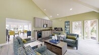 This 950-square-foot single-floor community space is thoughtfully designed to accommodate social gatherings and relaxation. The layout comprises a dining room, a combined kitchen and living room area, all unified by soaring vaulted ceilings that amplify the sense of openness throughout.

The dining room is bathed in natural light from large windows and glass doors that offer views of a pool and lush outdoor surroundings. Light beige tile flooring runs throughout the space, complementing the neutral gray walls. Round tables paired with chartreuse upholstered chairs provide flexible seating, while white decorative columns add an architectural elegance to the open floor plan.

The kitchenette is outfitted with taupe raised-panel cabinetry, granite countertops, an under-counter beverage refrigerator, a sink, and a dedicated coffee and beverage station — ideal for a community amenity space. Slate-toned tile flooring anchors this functional area.

The living room features soft green walls, vaulted ceilings, and a wall-mounted flat-screen TV set above a wood-toned media console. A tiled fireplace surround serves as a striking focal point. Plush blue-toned armchairs arranged on a striped area rug create an inviting lounge atmosphere. Glass doors connect the living area directly to the outdoors, ensuring abundant natural light throughout.