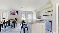 This single-floor community clubhouse is designed to offer residents a comfortable and functional shared gathering space. The open-plan layout seamlessly connects a kitchenette area with a casual dining and lounge zone, creating an inviting atmosphere for socializing.

The kitchenette is outfitted with a granite-topped island with beadboard paneling, white subway tile backsplash, open wood shelving with matte black hardware, a stainless steel mini-refrigerator, and a sink — all finished in a crisp, modern farmhouse style. Bar-height seating lines the island, making it ideal for casual dining or conversation.

The main lounge and dining area features light wood-look tile flooring, neutral walls, and multiple windows that bring in natural light and offer views of the surrounding grounds. A wall-mounted flat-screen television anchors the space, which is furnished with industrial-style round bistro tables and black metal chairs. A glass-paneled exterior door provides additional brightness and direct access to the outdoors.

A separate seating nook, carpeted with a bold teal and grey patterned area rug, provides a quieter retreat furnished with teal leather accent chairs and a small side table. Windows in this area overlook a pool and fenced outdoor recreation space. A restroom is also accessible from the main space, enhancing the functionality of this community facility.
