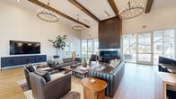Located at 4747 N 99th Ave in Phoenix, Arizona, this thoughtfully designed single-floor clubhouse offers a generous and open layout well-suited for gathering and relaxation. The main lounge area commands immediate attention with its soaring ceilings, exposed dark wood beam accents, and dramatic floor-to-ceiling slatted wood fireplace wall. A sleek linear gas fireplace serves as the room's focal point, framed by large floor-to-ceiling glass doors and windows that flood the interior with natural light and provide direct views to the outdoor patio area. Multiple seating arrangements featuring rich leather sofas and accent chairs are anchored by layered rugs and wood coffee tables, creating an inviting and sophisticated atmosphere. Elegant chandelier lighting adds warmth throughout the space. Light hardwood flooring flows continuously across the entire footprint, tying each zone together cohesively. A dedicated wet bar area features a dark granite-topped island with bar seating, flanked by open decorative shelving, a sink, and cabinetry finished in rich wood tones. The entry foyer welcomes visitors with a statement chandelier, bold art, and curated seating. Together, these elements create a modern yet warm communal space with a refined, upscale character.