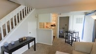 This two-floor space offers a practical and open layout that connects the living and kitchen areas seamlessly. The main level features wall-to-wall carpeting in the living area, which is bright and neutral in tone, creating a clean, welcoming atmosphere. A staircase with white-painted wood railings leads to the upper floor, positioned along one wall to maximize usable floor space.

The kitchen is defined by a half-wall partition that provides separation while maintaining an open connection to the living space. It is equipped with white cabinetry, a white refrigerator, and a range, giving it a clean, cohesive look. The white walls throughout the main level contribute to a light and airy feel.

Adjacent to the kitchen, a secondary room — visible through an open doorway — offers additional functional space. The living area accommodates a dining arrangement near a window that allows natural light to filter in. Overall, the space presents a straightforward, functional design with a neutral color palette, making it adaptable to a variety of furnishing styles and uses.