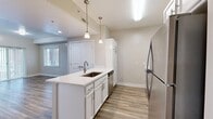 This efficiently designed single-floor unit offers a thoughtful layout across one bedroom, one bathroom, a combined kitchen and living area, and a hallway. The kitchen is a standout feature, showcasing a central island with a stainless steel undermount sink, crisp white cabinetry, light quartz countertops, and a full suite of stainless steel appliances including a refrigerator, range, and microwave. Pendant lighting above the island adds a modern touch, while wood-look flooring ties the kitchen and living spaces together seamlessly. The living area extends to sliding glass doors that open to an outdoor space, allowing natural light to fill the room. The bedroom is carpeted for comfort, features neutral warm tones, and includes a door with window access to natural light. The bathroom is clean and functional, appointed with a white vanity, undermount sink, mirror with contemporary lighting, and wood-look tile flooring that complements the rest of the unit. Throughout the space, a consistent neutral color palette, white trim, and modern fixtures create a cohesive and move-in-ready atmosphere.