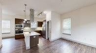 Located at 4700 Riverwood Cir in Raleigh, NC, this three-bedroom, three-bathroom multi-story home offers a thoughtfully organized layout across three floors. The ground level features a spacious two-car garage with a garage door opener, concrete flooring, and interior access to the main living areas. The main living floor showcases an open-concept design combining the kitchen, dining, and living areas, all finished with rich, dark hardwood-style flooring. The contemporary kitchen is a standout, featuring dark espresso cabinetry, a center island with a white countertop, a striking stainless steel overhead range hood, pendant lighting, stainless steel appliances including a French door refrigerator, double wall ovens, and a built-in microwave, all complemented by a light tile backsplash. Natural light flows in through multiple windows throughout. The upper floor hosts the bedrooms, including a generously sized primary bedroom, all carpeted or finished with hardwood-style flooring and ceiling fans for comfort. Bedrooms feature views of surrounding greenery through slatted blinds. Three full bathrooms serve the home, along with three closets for ample storage. A dedicated laundry area with a top-load washer adds convenience. The brick exterior and landscaped surroundings reflect a well-maintained community setting.