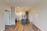 Located at 32 S Main St in downtown Concord, New Hampshire, this thoughtfully designed two-bedroom, one-bathroom residence offers a clean, contemporary aesthetic across a single floor. The open-concept living and kitchen area serves as the heart of the home, anchored by a striking kitchen island with a blue base and white quartz countertop, complemented by pendant lighting overhead. White shaker-style cabinetry, a marble-look tile backsplash, and a full suite of stainless steel appliances — including a refrigerator, range, microwave, and dishwasher — complete the well-appointed kitchen. Warm wood-style flooring flows throughout the main living areas, creating a cohesive and inviting atmosphere. A central hallway connects the bedrooms, closet, and bathroom, lined with clean white walls and louvered closet doors. The spacious primary bedroom offers natural light and comfortable proportions, while the second bedroom features a window with views of the surrounding neighborhood. The full bathroom is finished with wood-look tile flooring, a combined tub and shower, a wide vanity with warm-toned cabinetry, and a large mirror. Neutral gray tones and recessed lighting throughout lend the space a bright, modern feel.