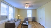 This single-floor space offers a clean, functional layout with a welcoming and understated aesthetic. The living area is a highlight of the space, featuring warm hardwood flooring that runs throughout and lends a sense of continuity and warmth. Multiple windows dressed with white blinds allow natural light to filter in generously, brightening the soft yellow-painted walls and creating an airy, open atmosphere. A ceiling fan with a built-in light fixture serves as a central focal point overhead, providing both practical ventilation and ambient lighting. The room comfortably accommodates a seating arrangement that includes a sofa and armchair, centered around a dark coffee table — ideal for relaxed lounging or casual gatherings. A classic cast-iron radiator positioned beneath one of the windows speaks to the building's traditional character and serves as a charming architectural detail. Crisp white trim and baseboards frame the room neatly, complementing the neutral color palette throughout. The overall ambiance is calm and straightforward, making it a practical and livable space with timeless design elements that offer a solid foundation for personalization.