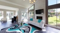 This single-story space makes an immediate impression with its dramatic vaulted ceilings and an abundance of natural light streaming through expansive windows and glass doors. The main living area serves as the focal point of the space, anchored by a floor-to-ceiling stacked stone fireplace wall with a mounted flat-screen television above. A tufted white Chesterfield sofa and bold teal accent pillows complement the grey-and-teal area rug, lending the space a contemporary yet inviting character. A chandelier overhead adds an elegant finishing touch.

Dark hardwood-style tile flooring flows consistently throughout, grounding the open layout and connecting the various zones seamlessly. A sunlit sitting area features large sliding glass doors and windows that overlook an outdoor pool, providing bright, airy views and easy outdoor access. White accent chairs and chrome side tables furnish this relaxed retreat.

The kitchen area is equipped with stainless steel appliances, including a full-size refrigerator and dishwasher, paired with light wood cabinetry and clean white countertops. Track lighting illuminates the workspace efficiently. A peninsula with white bar stools offers casual seating and defines the transition between the kitchen and the open living space beyond.
