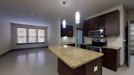This well-appointed single-floor unit offers a contemporary layout with a thoughtfully designed open-concept kitchen and living area. The kitchen is a standout feature, showcasing rich dark espresso cabinetry that extends from floor to ceiling, light granite countertops, and a full suite of stainless steel appliances including a gas range, built-in microwave, refrigerator with ice maker, and dishwasher. A subway tile backsplash adds a classic touch, while a central island with a rounded granite surface and bar-height seating provides additional workspace and dining functionality. Stylish pendant lighting illuminates the island from above, enhancing the kitchen's modern aesthetic. A dedicated pantry area offers generous storage space just off the main kitchen corridor. The open living area flows naturally from the kitchen and is bathed in natural light from a set of windows fitted with blinds. Smooth, neutral-toned flooring runs consistently throughout the unit, contributing to a clean, cohesive look. The bedroom is a comfortable, open room equipped with a ceiling fan and ample wall space. The bathroom features a soaking tub with tiled surround accented by dark decorative tile, a toilet, and a dark wood vanity with granite countertop and integrated storage. Overall, the unit presents a polished, modern interior with consistent finishes throughout.