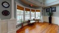 Located at 5806 Ogeechee Rd in Savannah, Georgia, this single-story clubhouse facility combines Southern charm with refined interior design throughout its generously proportioned open layout. Warm pine hardwood floors flow seamlessly across the entire space, anchoring each area with natural character and continuity.

The main gathering area serves as the heart of the space, centered around a striking full-height exposed brick fireplace featuring a rustic reclaimed wood mantel. Decorative white wainscoting, elegant columns, and crown molding frame the room, lending a classic architectural sophistication. A sun-drenched bay window area with tall, grid-paned windows and woven Roman shades offers an inviting reading nook with sweeping views of the outdoor pool and lush landscaping beyond.

The fully equipped kitchen is outfitted with rich dark cherry cabinetry extending to the ceiling, dark granite countertops with a mosaic tile backsplash, and stainless steel appliances including a wall oven and side-by-side refrigerator. Recessed lighting illuminates the space efficiently. An adjacent casual dining nook with a round bistro table and bar-height seating provides a comfortable spot for informal gatherings.

The open-concept doll's-eye view reveals a spacious, well-connected floor plan with defined zones for lounging, dining, and socializing, all unified by consistent finishes and graceful architectural detailing throughout.
