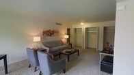 This single-floor space presents a cozy and functional living area finished with wall-to-wall carpeting that adds warmth underfoot. The main seating area is anchored by a tufted gray sofa accompanied by two matching armchairs, all arranged around a dark-stained rectangular coffee table — creating an inviting, conversational layout. A decorative canvas featuring an autumn tree in warm reds and oranges adds a focal point of color to the neutral walls.

Ambient lighting is provided by two table lamps that cast a soft, warm glow throughout the room, complementing the flush-mount ceiling fixture. A compact shelving unit tucked into the corner offers display and storage space. Ventilation is handled through a ceiling-mounted HVAC vent, pointing to a climate-controlled interior.

A standout feature of this space is the generous storage provided by two side-by-side closets along the far wall, both equipped with interior shelving and hanging rods — ideal for organized storage. The overall ambiance is understated and practical, with neutral tones throughout that lend the space a clean, versatile character suited to a variety of uses.