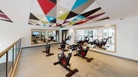 Located at 1055 W Town and Country Rd in Orange, California, this contemporary amenity space is a single-floor facility that combines energetic design with thoughtful functionality. The space is immediately distinguished by its striking geometric ceiling panels, featuring bold triangular shapes in a vivid palette of teal, red, purple, chartreuse, and charcoal — an artistic design element that carries consistently throughout the space and gives it a dynamic, modern identity.

The spin studio is a standout feature, equipped with a fleet of Peloton cycling bikes arranged for group fitness sessions. Full-length mirrors line the walls, amplifying the sense of space and allowing riders to monitor their form. The same colorful geometric ceiling treatment crowns this room, reinforcing the cohesive design language found throughout.

A light-filled corridor or mezzanine-level overlook provides sweeping views of a resort-style courtyard below, complete with a pool, lounge seating, and lush landscaping framed by a modern multi-story residential building. Large black-framed floor-to-ceiling windows flood the area with natural light, while sculptural globe pendant chandeliers on brass fixtures add a warm, sophisticated glow. A warm wood handrail lines the open balustrade, completing the upscale, welcoming ambiance of this well-appointed amenity space.