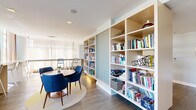 Located at 160 Morgan Street in Jersey City, NJ, this single-floor communal living space offers a well-appointed and versatile environment designed for both relaxation and productivity. The open layout is anchored by wide-plank hardwood flooring and bright white ceilings with recessed lighting, creating a clean and airy aesthetic throughout.

A defining architectural feature is the floor-to-ceiling built-in bookshelf unit that serves as a natural room divider, organizing the space into distinct functional zones while maintaining an open, connected feel. One side of the divider hosts a cozy lounge area furnished with plush gray armchairs, ideal for quiet reading or casual conversation. On the other side, a round dining table surrounded by navy blue upholstered chairs sits atop a circular rug, providing an inviting gathering spot.

A long, built-in counter-height workspace runs along the window-facing wall, lined with bar stools and bathed in natural light streaming through floor-to-ceiling windows dressed with sheer curtains. This area is well-suited for individual work or study. Soft sage green accent walls complement the neutral palette, while curated wall art adds a touch of character. The overall ambiance is calm, modern, and welcoming — a refined communal retreat.