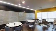 This contemporary single-floor workspace is thoughtfully designed to foster collaboration and creativity. The interior makes a strong visual statement with a striking combination of dark accent walls, bold yellow accents, and rich hardwood flooring that grounds the space with warmth.

A standout feature is the expansive glass whiteboard wall that spans the length of one side of the room, offering an ideal surface for brainstorming, ideation, and team planning sessions. The wall's transparent design adds depth while maintaining an open, airy atmosphere.

The seating arrangement includes multiple round marble-top tables paired with sleek, modern chairs in a deep charcoal upholstery, creating intimate groupings suited for small team discussions or informal meetings. A lounge-style seating area near the large windows adds a relaxed alternative workspace, bathed in natural light that brightens the room.

Overhead, a mix of recessed lighting and pendant track fixtures provide layered illumination, complemented by a continuous LED strip light that adds a refined architectural detail along the ceiling's edge. The overall ambiance is polished yet approachable — a well-equipped environment designed to support dynamic, collaborative work.
