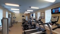 This single-floor fitness center offers a comprehensive workout environment designed to accommodate a range of exercise needs. The space is outfitted with multiple cardio machines, including treadmills and elliptical trainers, positioned to take advantage of the natural light streaming in through the large windows fitted with white blinds. Strength training equipment lines the perimeter, providing users with a well-rounded selection of workout options. The room features warm yellow walls accented by white crown molding and baseboards, lending a clean and polished aesthetic. Overhead, a series of fluorescent light fixtures ensure the space is brightly and evenly illuminated throughout. A wall-mounted television is positioned centrally, allowing users to stay entertained during their workouts. Motivational fitness posters decorate the walls, contributing to an energizing atmosphere. A wall clock and an emergency exit sign are also visible, reflecting attention to both functionality and safety. The flooring is carpeted with exercise mats placed beneath the equipment for added comfort and protection. An interior door leads to an adjoining area, enhancing the overall flow of the facility.