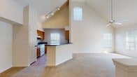 This single-floor residence makes a striking first impression with its dramatic vaulted ceilings that soar over the open-concept living and kitchen area, creating an expansive, airy atmosphere. Track lighting illuminates the kitchen, which features warm wood cabinetry, dark countertops, a peninsula breakfast bar, and tile flooring. White appliances, including a refrigerator and range, are complemented by natural light streaming through a window above the sink area. The living space flows seamlessly from the kitchen, showcasing plush carpeting and multiple windows fitted with blinds that bathe the room in soft, natural light. A ceiling fan adds comfort to the generous living area. The bedrooms are carpeted and well-proportioned, each equipped with ceiling fans and ample closet storage via bi-fold doors with built-in hanging rods. The bathrooms are finished with warm-toned tile flooring, wood vanities with granite-style countertops, large mirrors, and a tub-shower combination. One bathroom features a walk-in closet accessible directly from the bath, adding a practical touch. Throughout the residence, neutral tones and consistent finishes create a cohesive, welcoming environment.