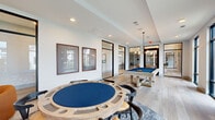 This single-floor game room offers a well-appointed recreational space designed for social engagement and leisure. The layout is thoughtfully organized to accommodate multiple activities simultaneously without feeling congested. At the center of the room, a substantial round poker table with a blue felt surface and carved wooden surround serves as a primary focal point, surrounded by matching game chairs atop a circular area rug. A full-size pool table with a blue felt top is positioned toward the opposite end of the room, suspended beneath a pendant light fixture that highlights the playing surface.

Along one wall, a built-in banquette seating area flanks a textured stone accent wall with a wall-mounted flat-screen television, complemented by warm pendant lighting and deep navy accent panels. Framed game-themed artwork lines the adjacent wall, adding a curated aesthetic touch. Lounge seating, including a rounded cognac accent chair and side table, provides casual gathering options near the floor-to-ceiling black-framed windows, which introduce generous natural light and direct views of the surrounding landscaping. Light-toned wide-plank flooring runs throughout, enhancing the bright, contemporary atmosphere. Black-framed glass partitions connect this space to an adjoining amenity area.