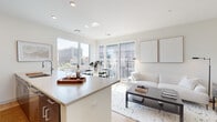 Located at 10 Commerce Dr in New Rochelle, NY, this modern single-floor residence offers a thoughtfully designed open-concept layout finished with light hardwood flooring and crisp white walls throughout. The kitchen is a standout feature, equipped with stainless steel appliances including a refrigerator, microwave, and range, complemented by sleek taupe cabinetry, a tile backsplash, and a generous island with a quartz countertop that comfortably accommodates bar seating — ideal for casual dining and entertaining. The kitchen flows seamlessly into the bright living area, where floor-to-ceiling windows and sliding glass doors invite abundant natural light and provide direct access to an outdoor balcony with urban views of the New Rochelle skyline. The primary bedroom is generously sized with large windows framing city views, while a secondary bedroom offers a cozy, well-appointed retreat with closet storage. Two bathrooms serve the residence, each featuring large-format tile flooring, clean-lined vanities with slate-blue cabinetry, and tiled shower surrounds. A well-proportioned hallway connects the private areas of the residence, adding to the functional and organized flow of the space. Recessed lighting throughout enhances the clean, contemporary aesthetic.
