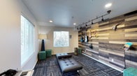 This single-floor space is designed as a dedicated music lounge, blending functional design with creative character. The room's most prominent feature is a full-height accent wall clad in horizontal planks of reclaimed wood in varied tones of grey, brown, and beige, giving the space a rustic yet contemporary aesthetic. Mounted along this wall is a curated collection of string instruments — including guitars, a ukulele, and a mandolin — displayed as both functional pieces and decorative art.

A track lighting system runs across the ceiling above the accent wall, providing directional illumination to highlight the instruments and enhance the room's visual warmth. Recessed ceiling lights throughout the space ensure even, ambient lighting overall. Natural light enters through a window on the opposite wall, lending the room a bright and open feel during daytime hours.

The flooring consists of patterned carpet tiles in a multi-tonal grey palette, adding texture underfoot while contributing to the room's acoustic character. Seating includes low-profile tufted leather benches and compact green upholstered cubes, creating a casual, inviting atmosphere well-suited for listening, practicing, or informal gatherings. A tall floor lamp in the corner adds a warm supplementary light source.