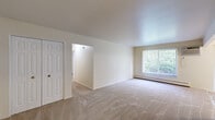 This single-floor unit offers a clean, functional layout designed for comfortable everyday living. The living area is generously proportioned, featuring neutral-toned wall-to-wall carpeting, a large window with vertical blinds that floods the space with natural light and frames views of surrounding greenery, and a baseboard heating system. A hallway connects the living area to the rest of the unit, providing a natural flow between spaces.

The galley-style kitchen is efficiently designed, featuring white cabinetry with ample storage above and below, light-toned countertops, a stainless single-basin sink, and white appliances including an electric range and a full-size refrigerator. The tile-style flooring adds a practical, easy-to-maintain surface underfoot.

The bedrooms are carpeted and finished in soft, neutral tones, each equipped with windows overlooking lush outdoor greenery and baseboard heaters for year-round comfort. One bedroom includes a closet with a classic paneled door, offering practical storage. The overall palette of warm whites and light beiges creates a calm, cohesive ambiance throughout the unit. The layout, as seen from the overhead floorplan, is compact yet well-organized, making efficient use of every area within the space.
