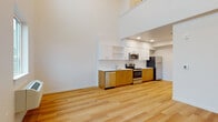 This contemporary two-story residence is defined by its dramatic double-height ceilings in the main living area, which create an expansive, airy atmosphere flooded with natural light from large windows and elevated clerestory panes. The open-concept layout seamlessly connects the living space to a well-appointed kitchen featuring warm wood-toned lower cabinetry, crisp white upper cabinets with open shelving, white countertops, and a full suite of stainless steel appliances including a range, over-the-range microwave, dishwasher, and refrigerator. Light hardwood-style flooring runs throughout the main level, enhancing the bright, clean aesthetic. A staircase leads to the upper level, where a loft-style bedroom area is carpeted for comfort and features a spacious walk-in closet and an open railing overlooking the living space below. An additional bedroom on the main level is also carpeted and includes closet storage. The bathroom is finished with a wood-toned vanity, white countertop, undermount sink, and a full tub-shower combination. Clean white walls, recessed lighting, and a consistent modern design language unify both levels into a cohesive, move-in-ready living environment.